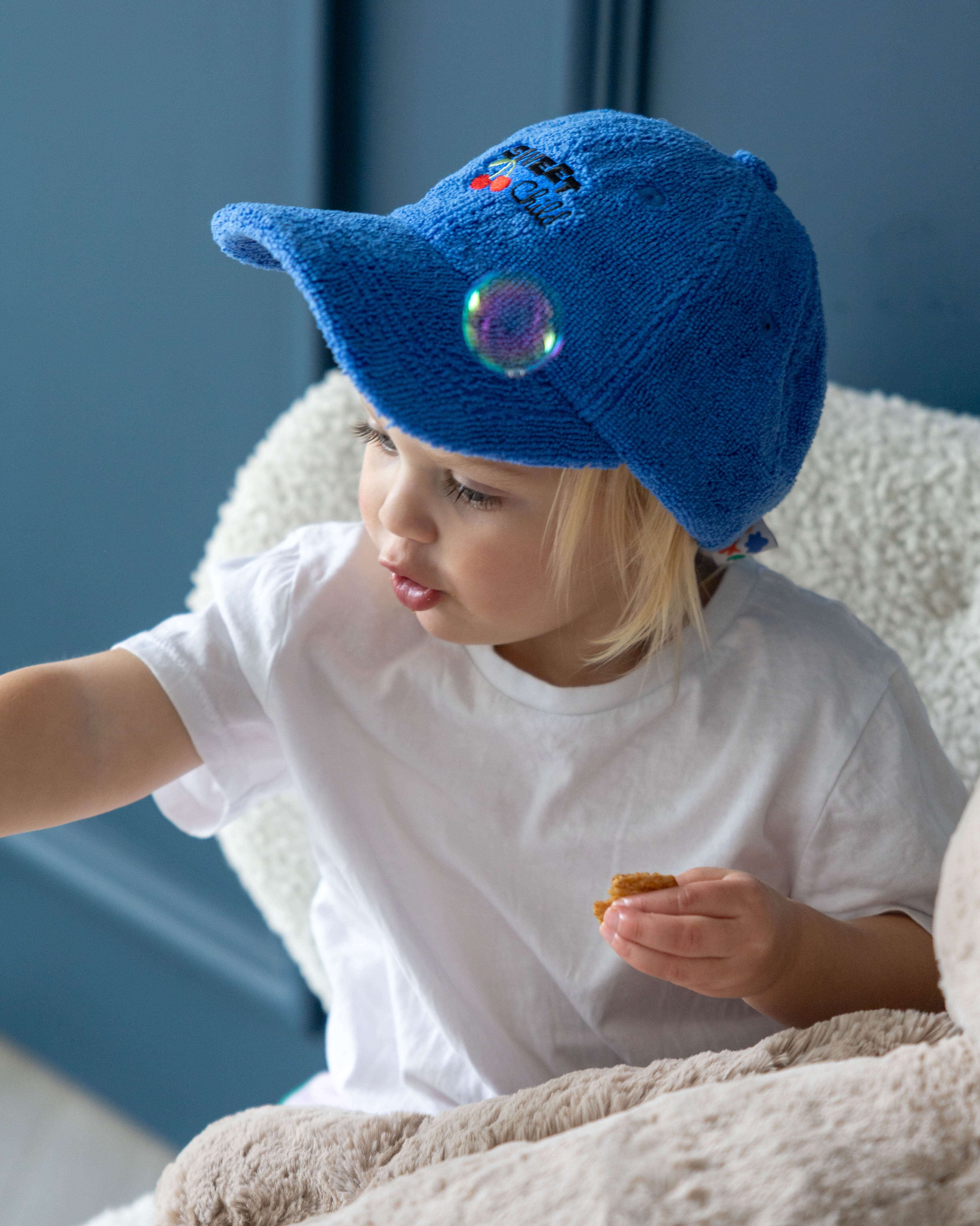 Child wearing a blue terry towelling hat with a 'sweet child' embroidery design, sitting on a couch.