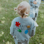 Child wearing a light blue sweatshirt with colorful designs and text in an outdoor setting.