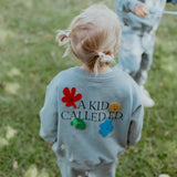 Child wearing a light blue sweatshirt with colorful designs and text in an outdoor setting.