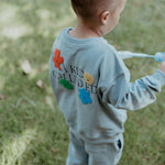 Child wearing a light blue tracksuit with colorful patches and text, standing on grass.