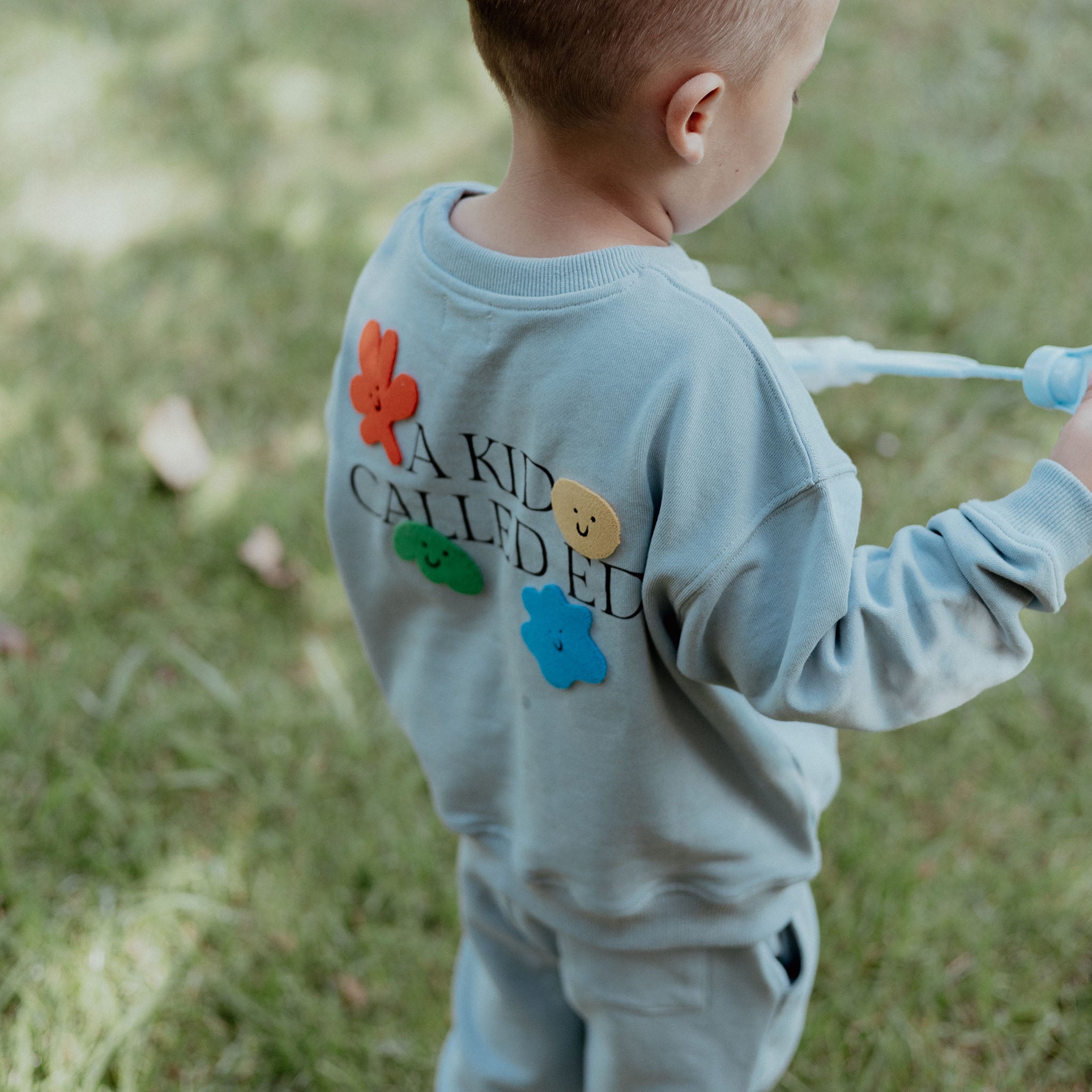 Child wearing a light blue tracksuit with colorful patches and text, standing on grass.