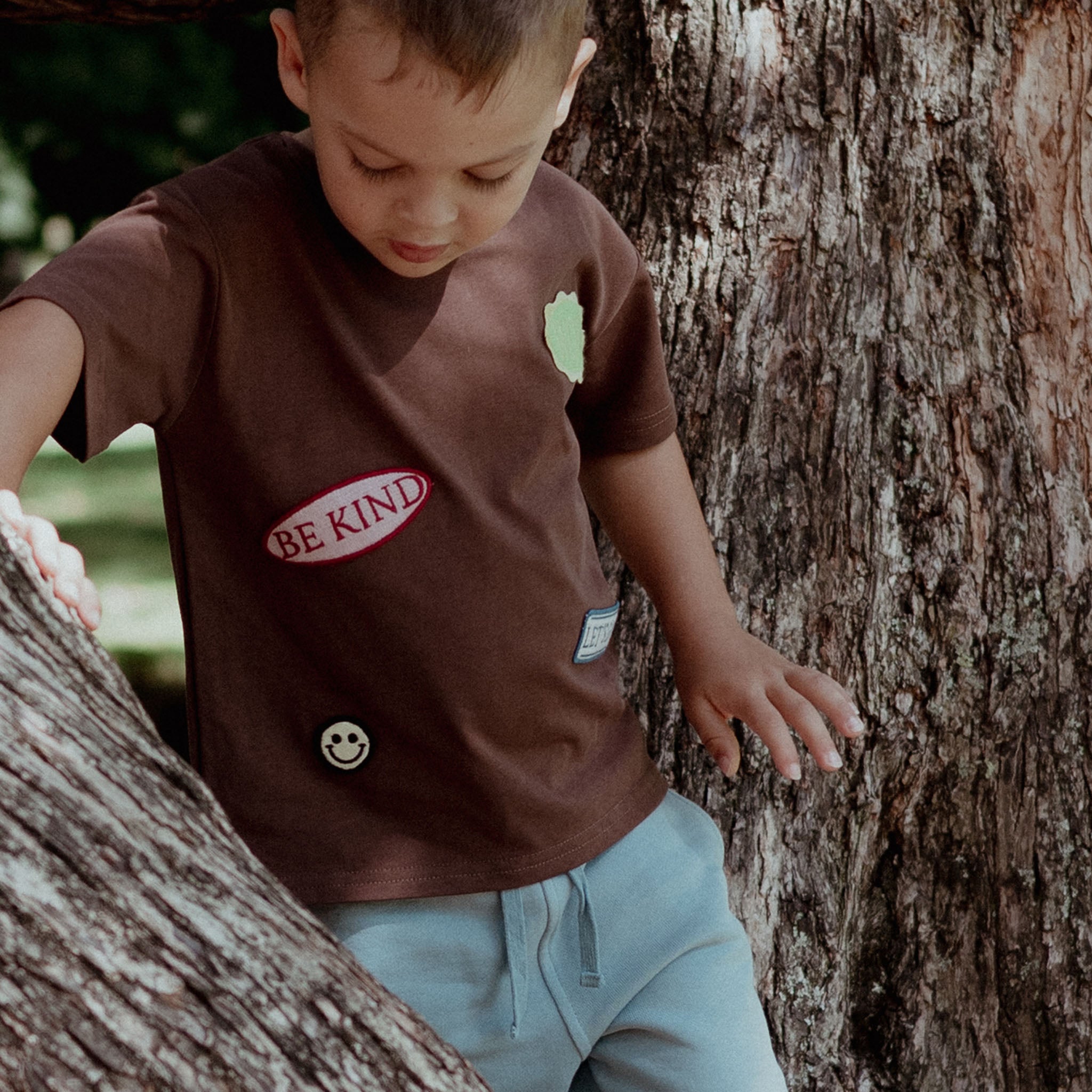 Child wearing a 'Be Kind' t-shirt standing next to a tree