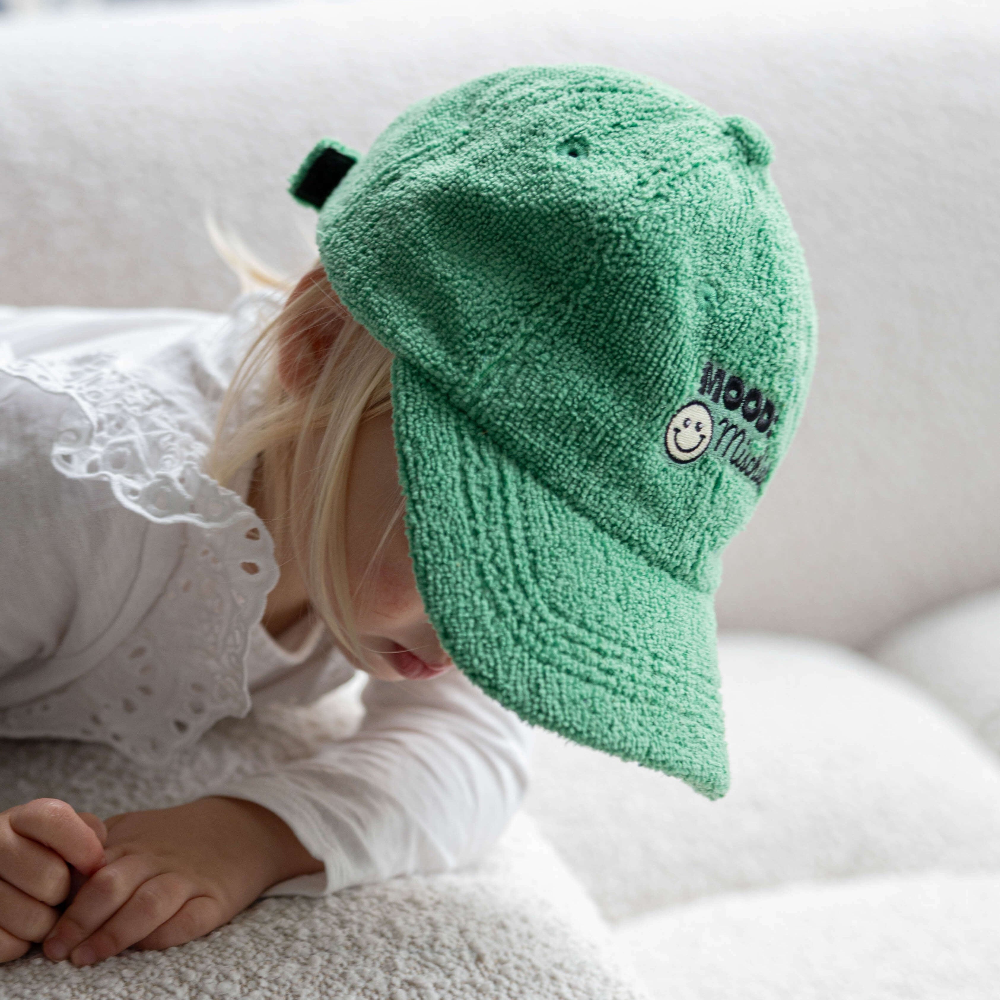 Child wearing a green cap with a smiley face logo on a white couch
