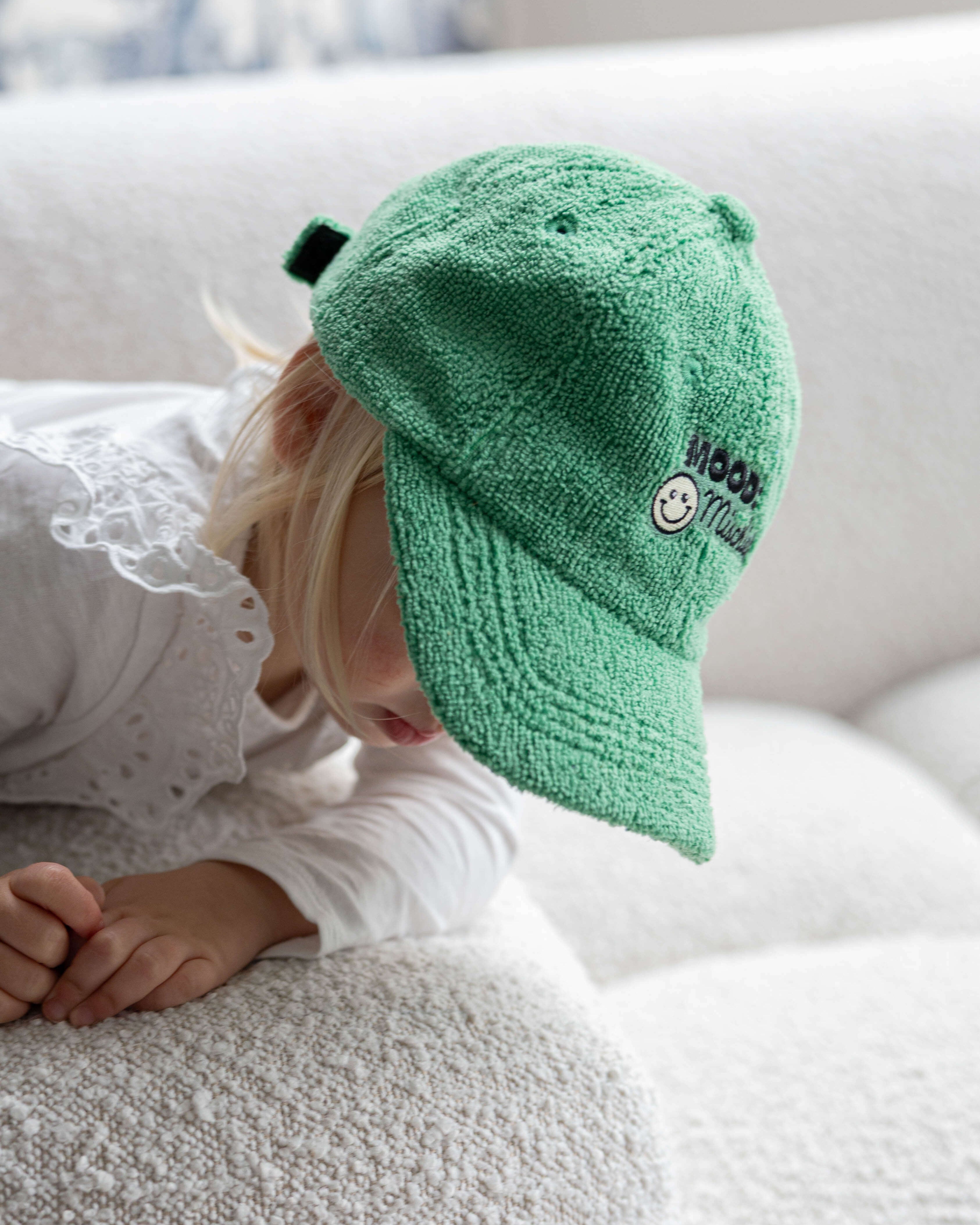 Child wearing a green cap with a smiley face logo on a white couch