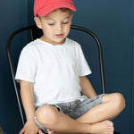 Child sitting on a black stool wearing a red cap and white shirt against a dark blue wall.
