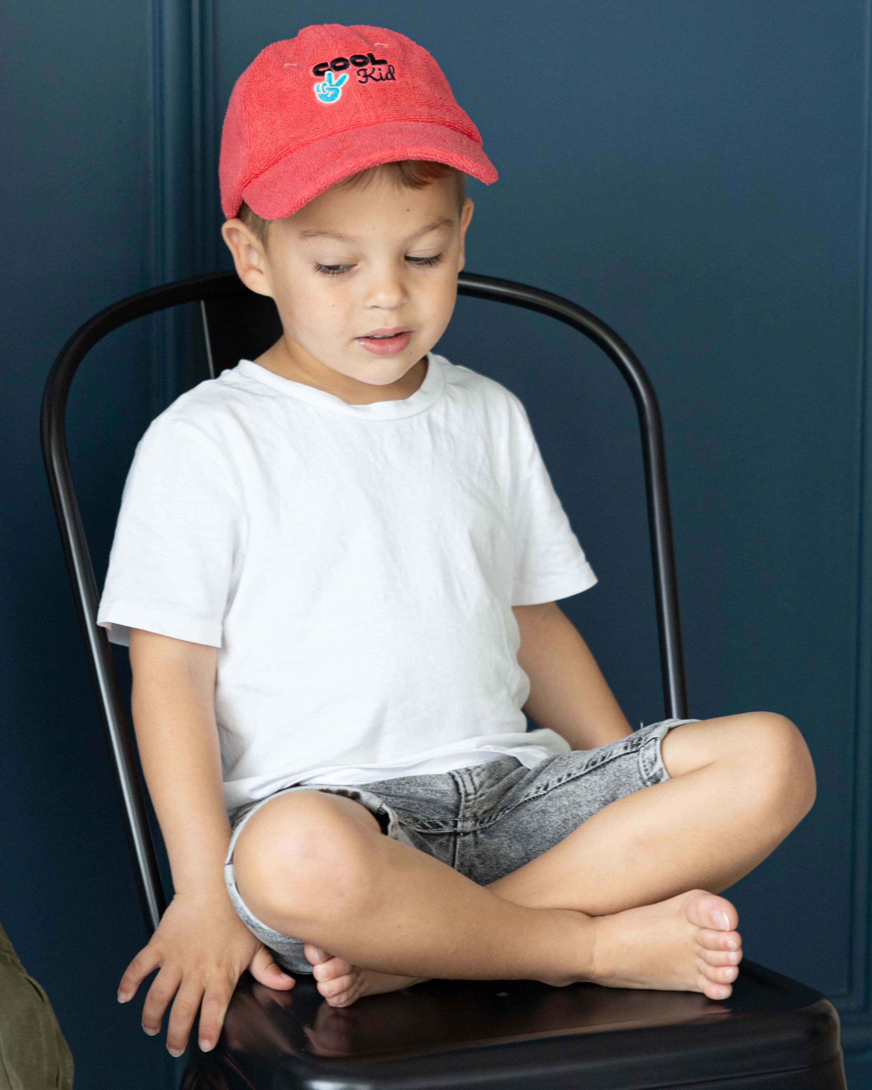 Child sitting on a black stool wearing a red cap and white shirt against a dark blue wall.