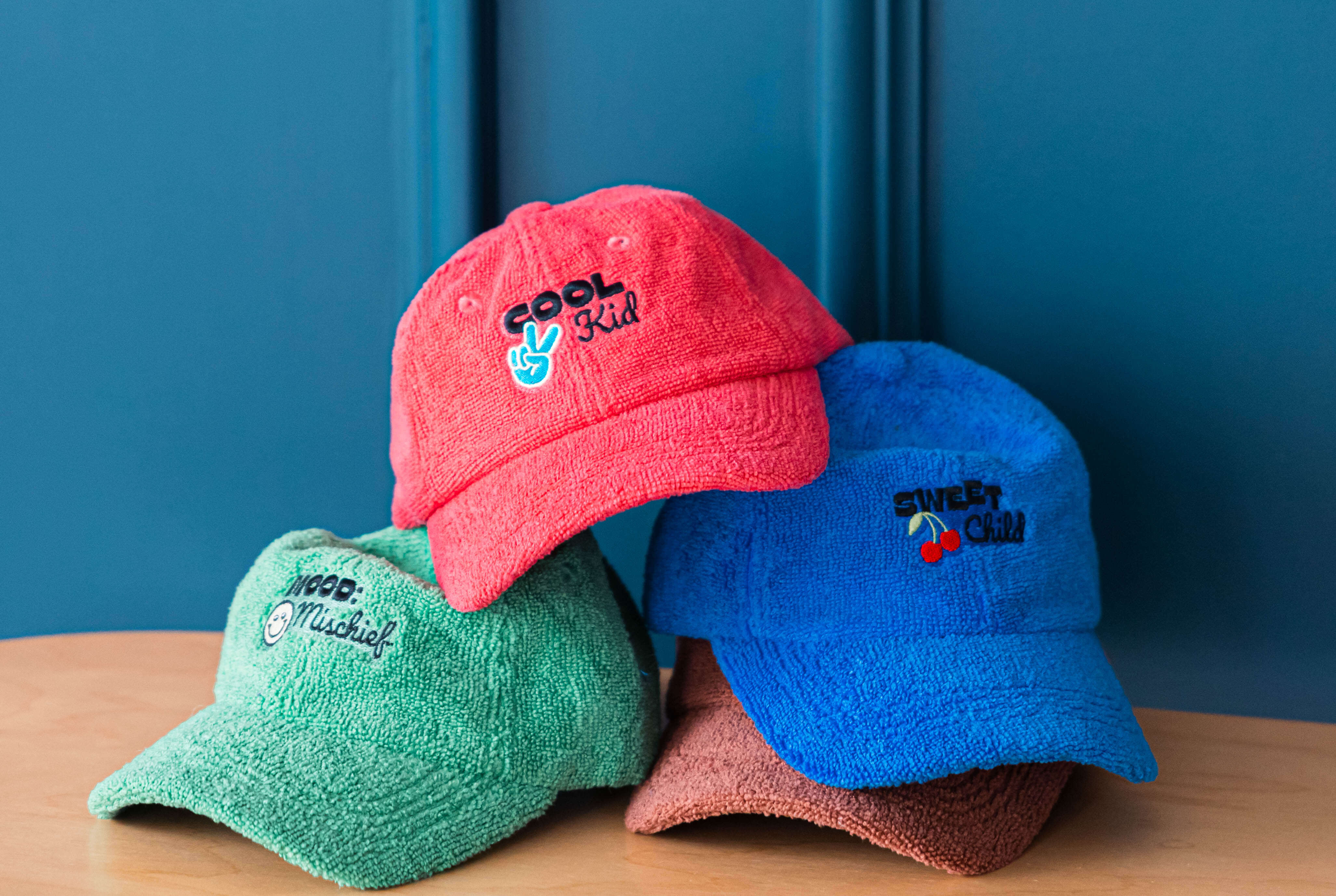 Four colorful caps with embroidered text on a wooden surface against a blue wall.