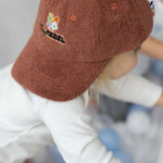 Child wearing a brown terry towelling cap with embroidery, standing in a playground.