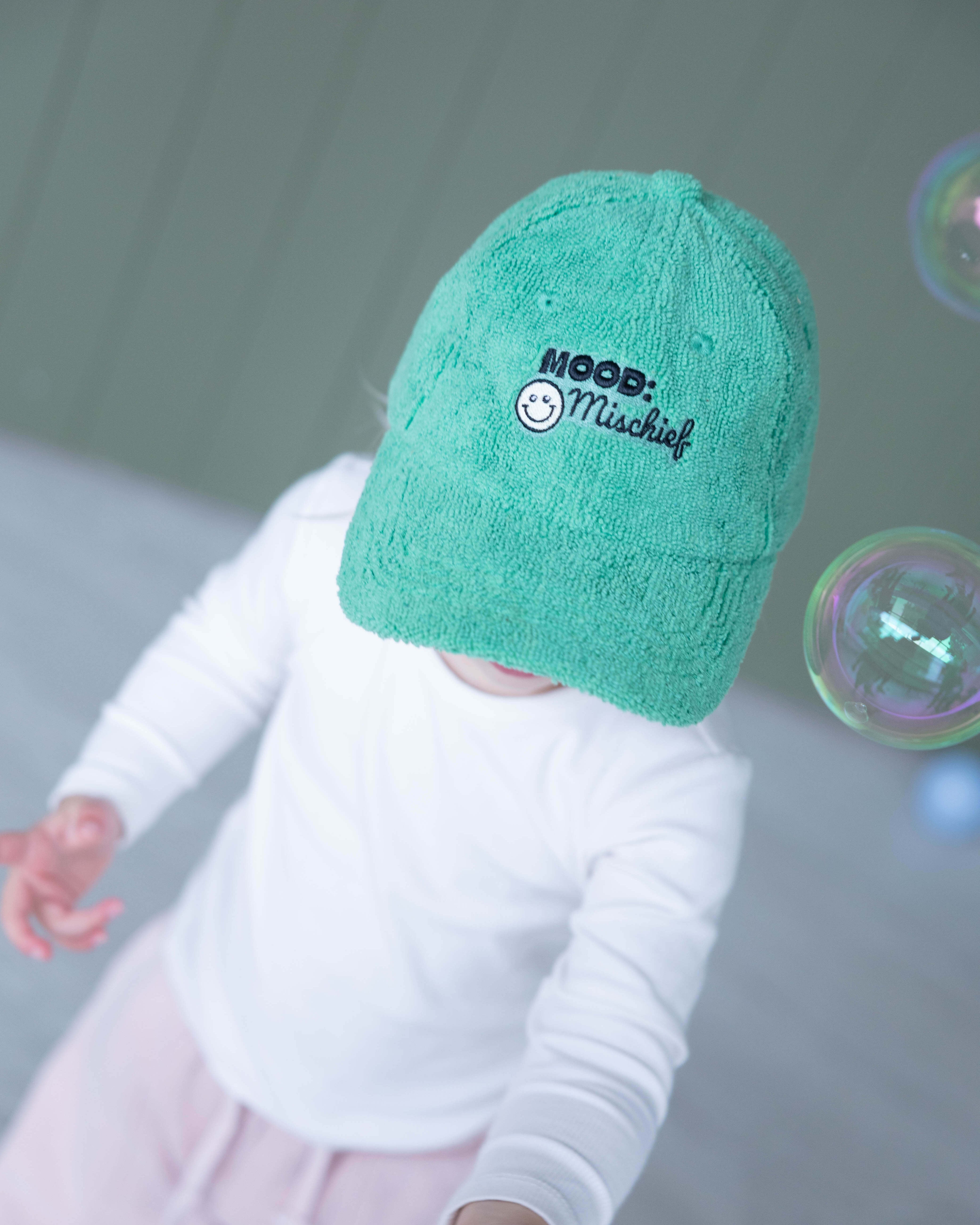 Child wearing a green towel with 'Mood: Mischief' text, surrounded by bubbles on a gray background