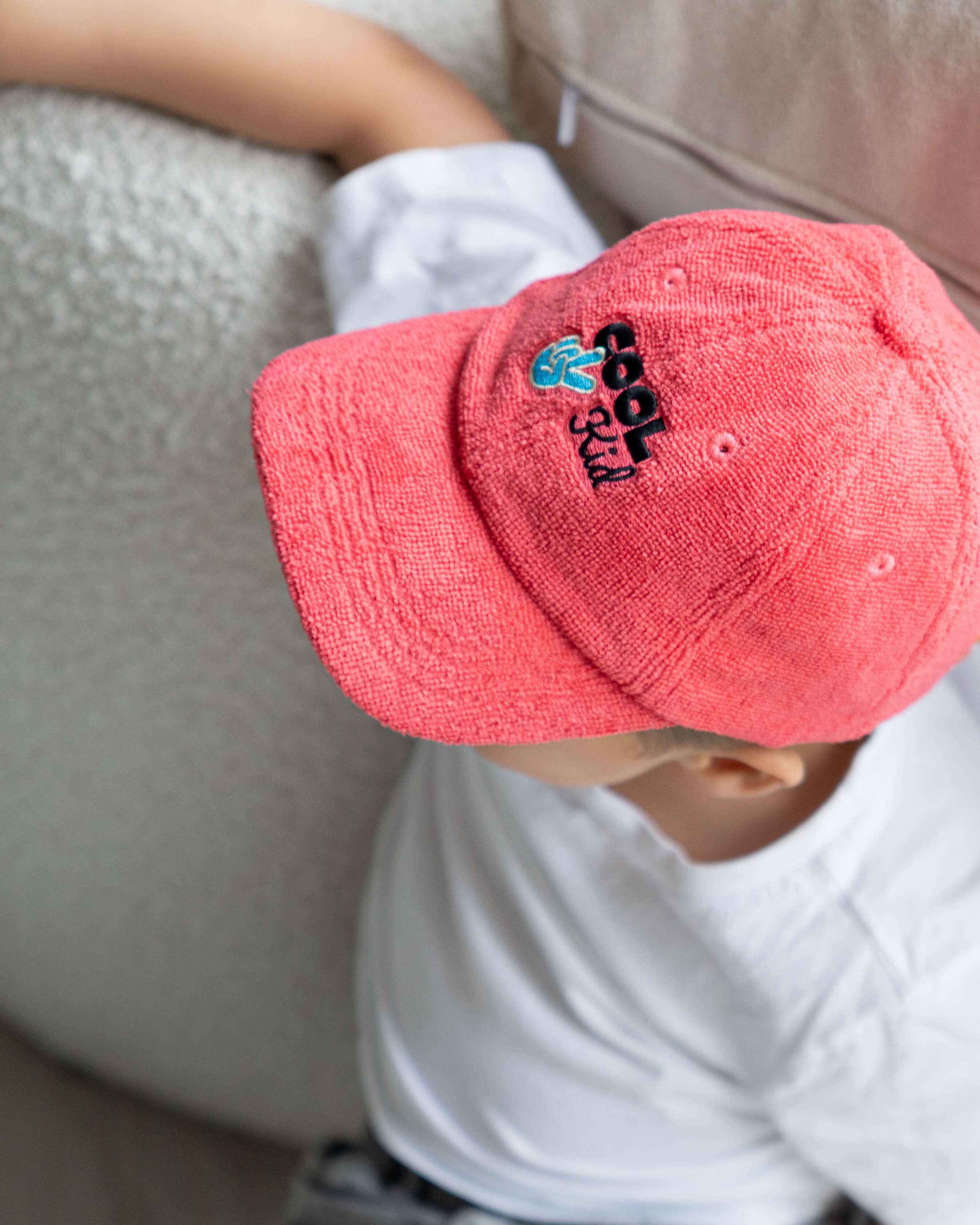 Child wearing a red cap with a cool kid logo, sitting on a couch.
