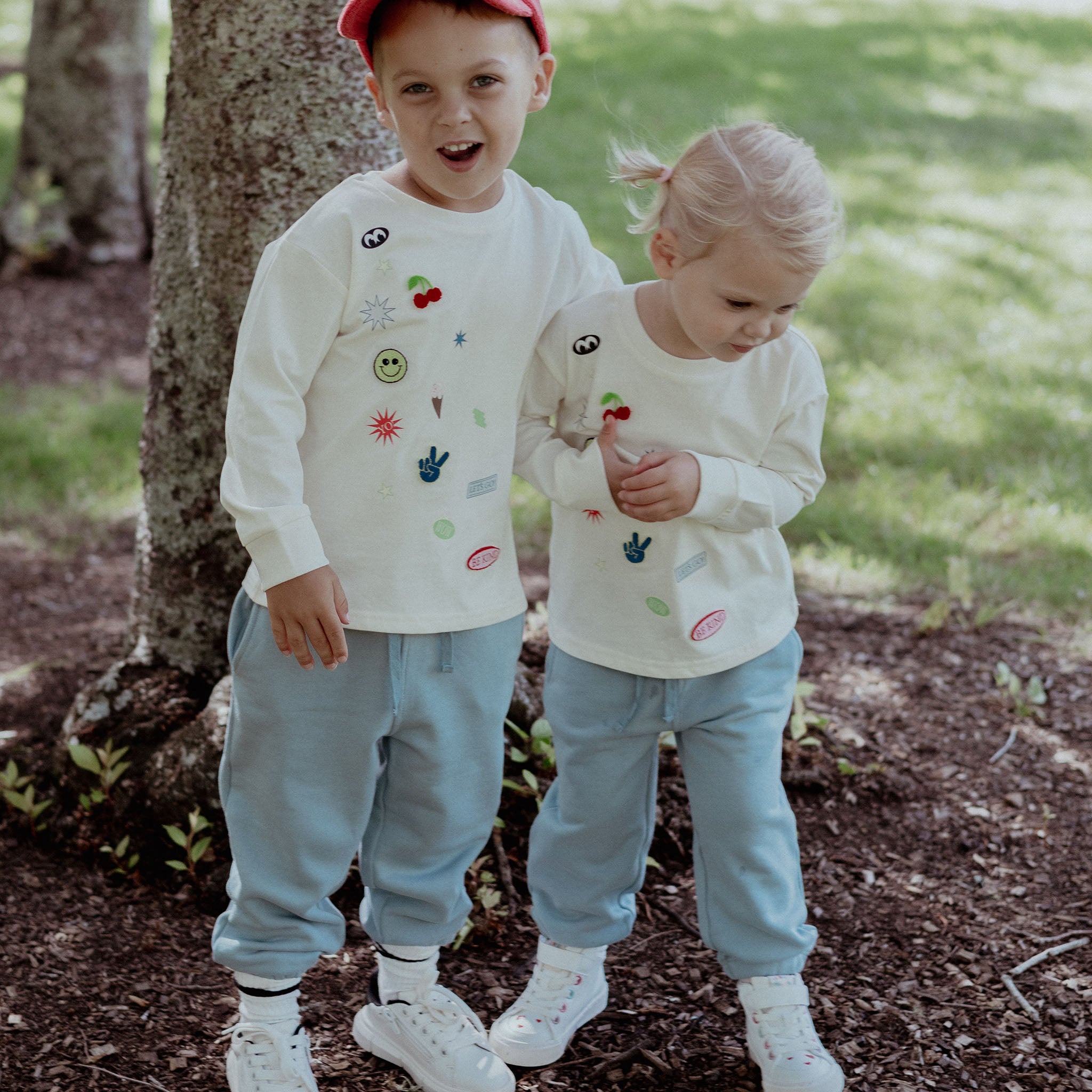 Two children wearing matching outfits with colorful designs standing outdoors near a tree.
