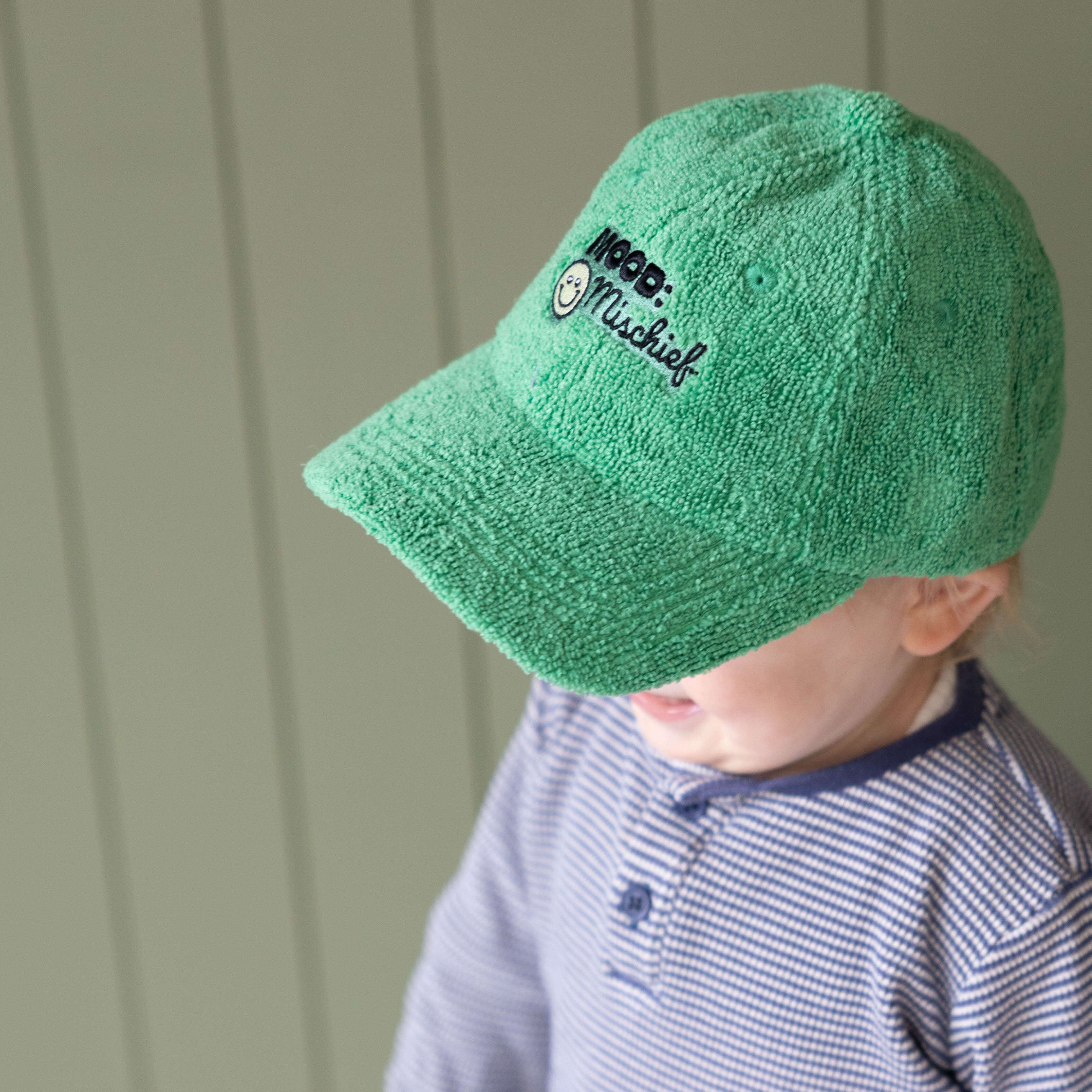 Child wearing a green cap with a logo against a light green wall.