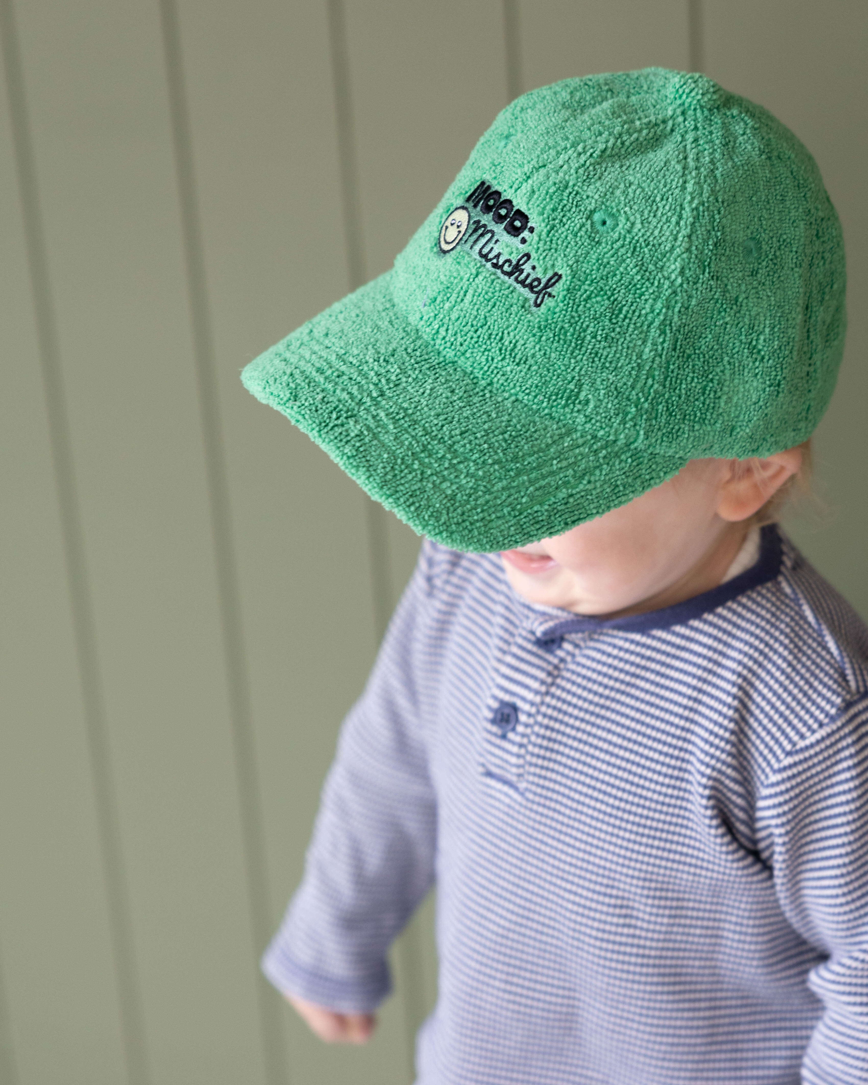 Child wearing a green cap with a logo against a light green wall.