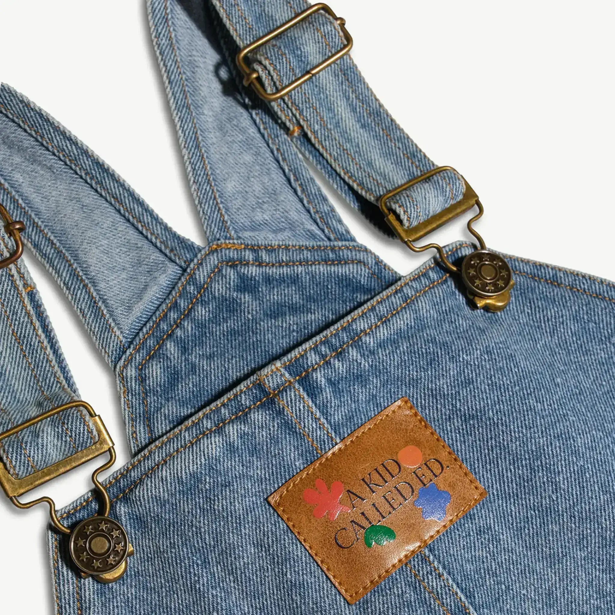 Close-up of the adjustable strap and leather logo patch on the kids denim overalls