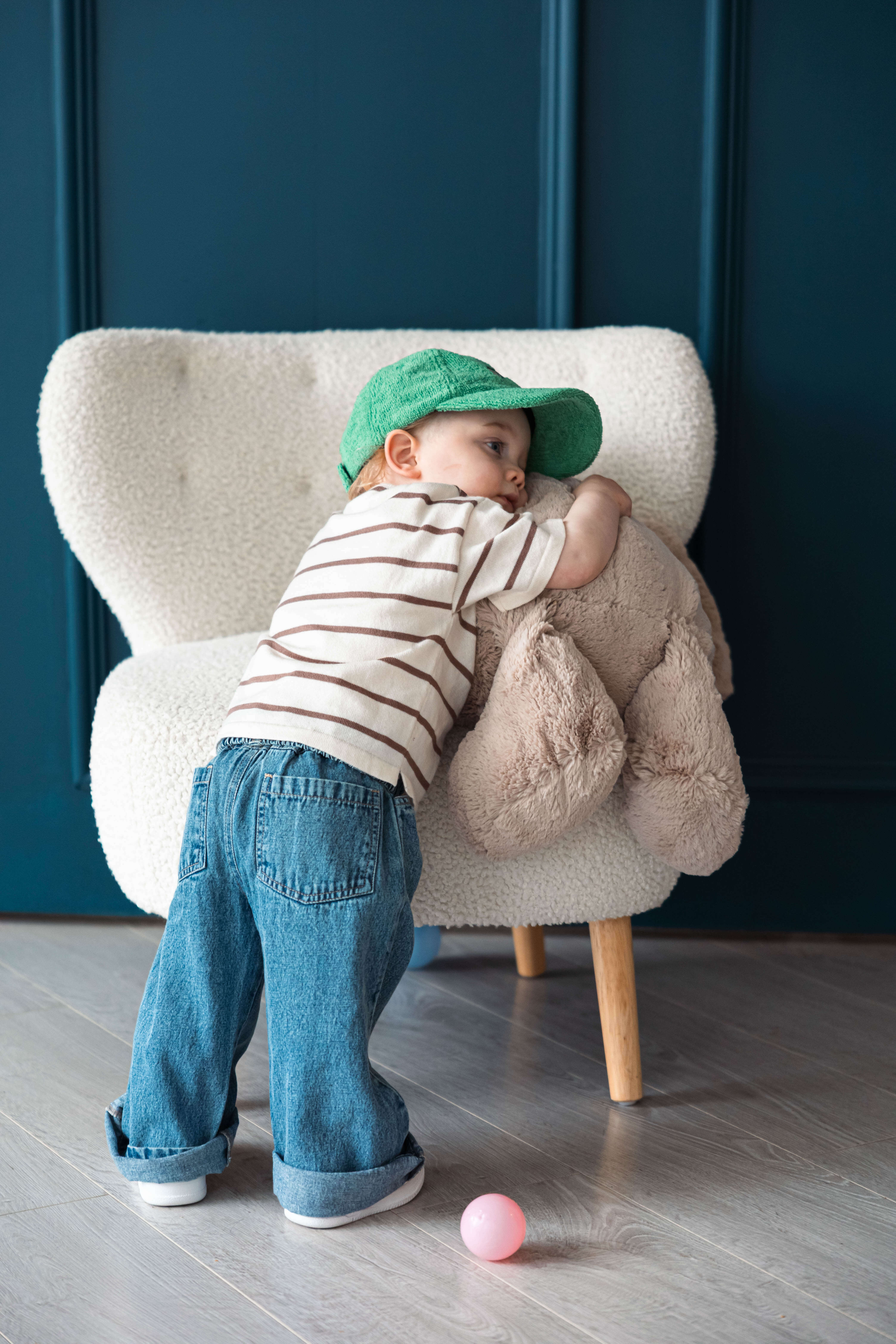Child wearing a green cap and striped shirt, standing next to a plush toy on a chair.