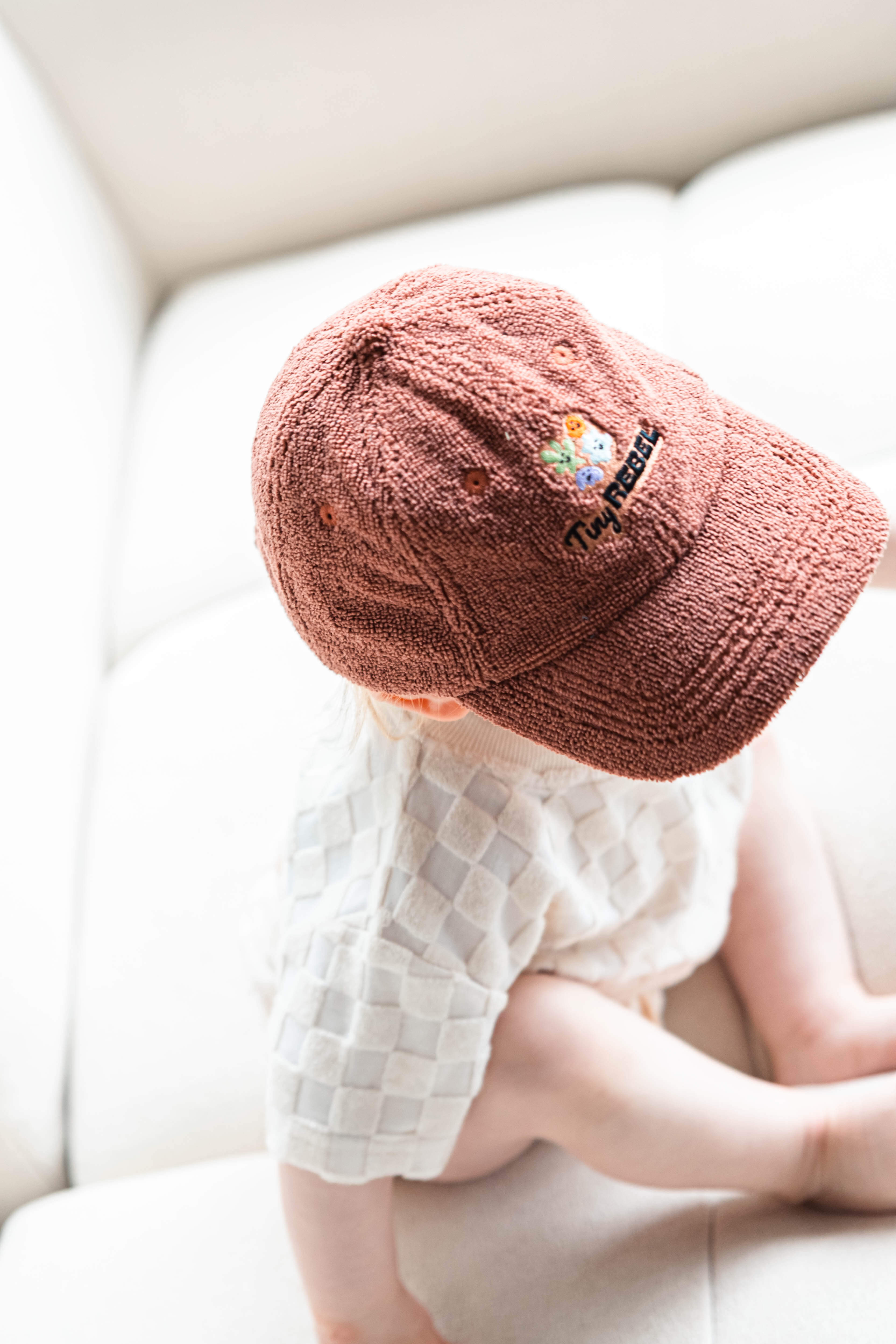 Toddler wearing a brown terry towelling embroidered cap on a white couch
